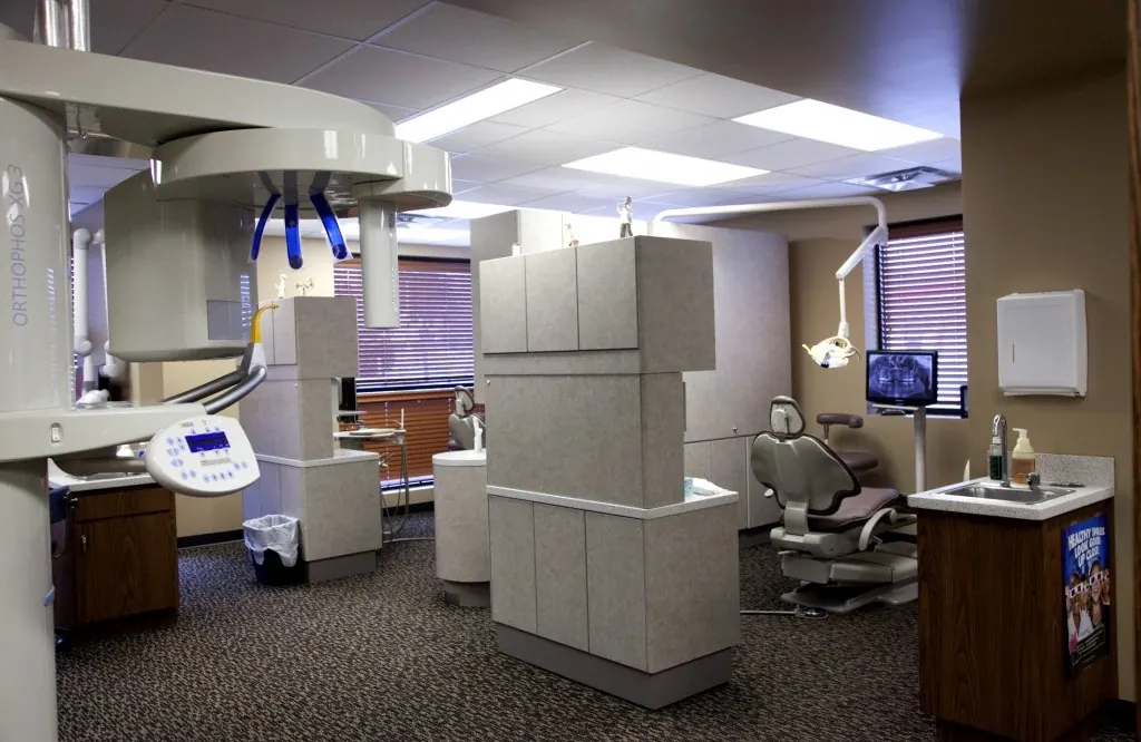 A treatment room with a patient chair, an overhead monitor and a panoramic imaging machine.