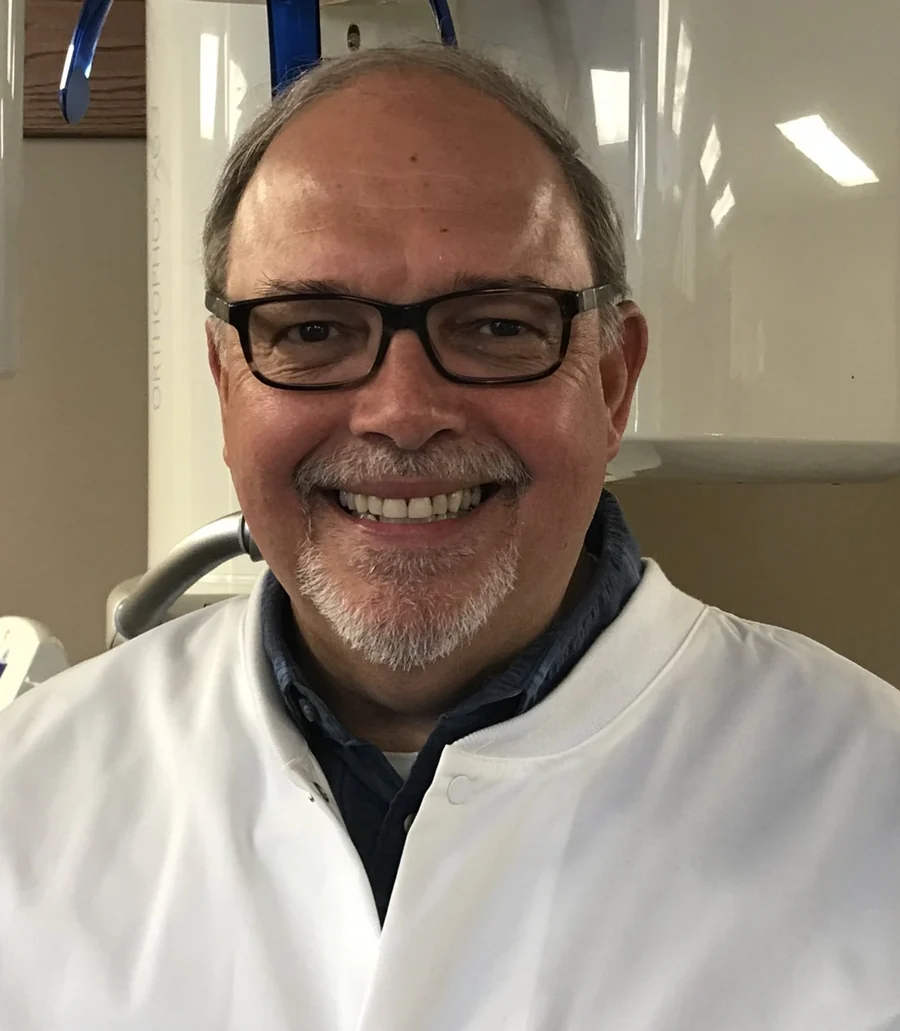 Dr. Terry Bolamperti, smiling, in glasses and a white clinic jacket, in a treatment room.